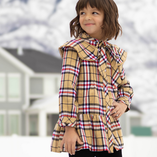 Child wearing the Checkered Charm Dress in tan, black, white, and red plaid, with a lace-trimmed collar and neck bow.
