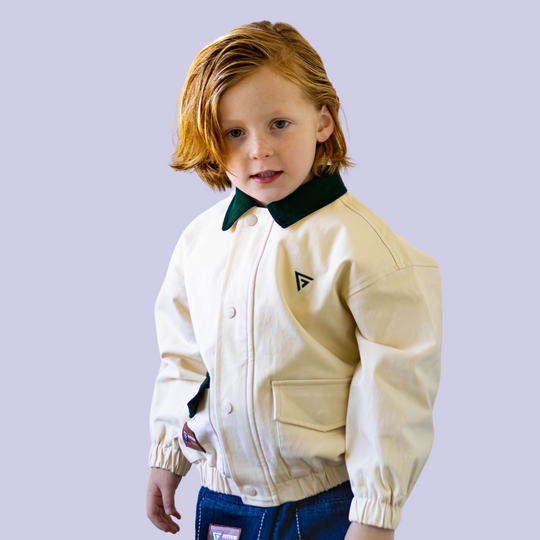 Cream Cruiser Jacket modeled on a child, showing its cream body, dark green collar, flap pockets, and black chest logo.