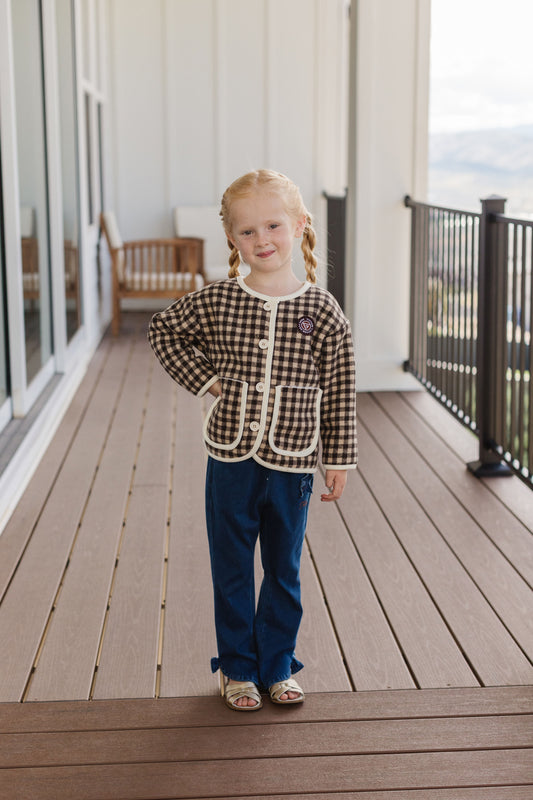 A child wears the Checkered Cub Jacket with brown-and-cream checks, white piping and front pockets on a porch.