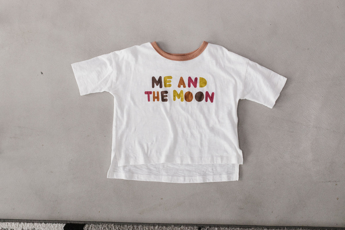 The white Luna Graphic Tee is shown flat with a brown neckline, colorful Me and the Moon lettering and a longer back hem.
