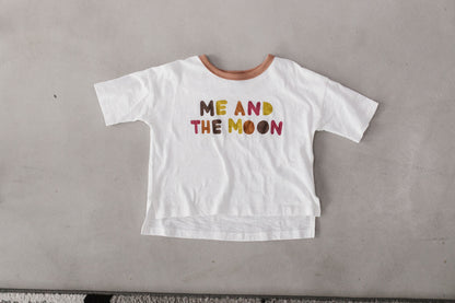 The white Luna Graphic Tee is shown flat with a brown neckline, colorful Me and the Moon lettering and a longer back hem.