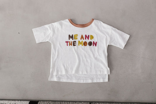 The white Luna Tee is shown flat with a brown neckline, colorful Me and the Moon lettering and a longer back hem.