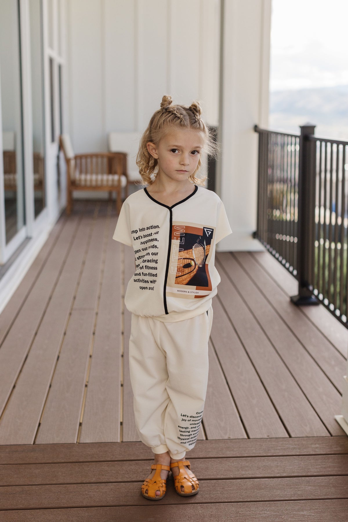 A child wears The Nori Set with an ivory graphic shirt and matching pants, black trim and tennis imagery.