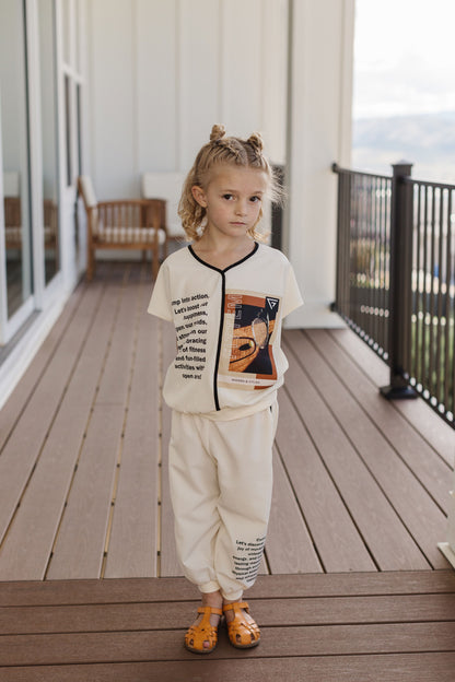 A child wears The Nori Set with an ivory graphic shirt and matching pants, black trim and tennis imagery.